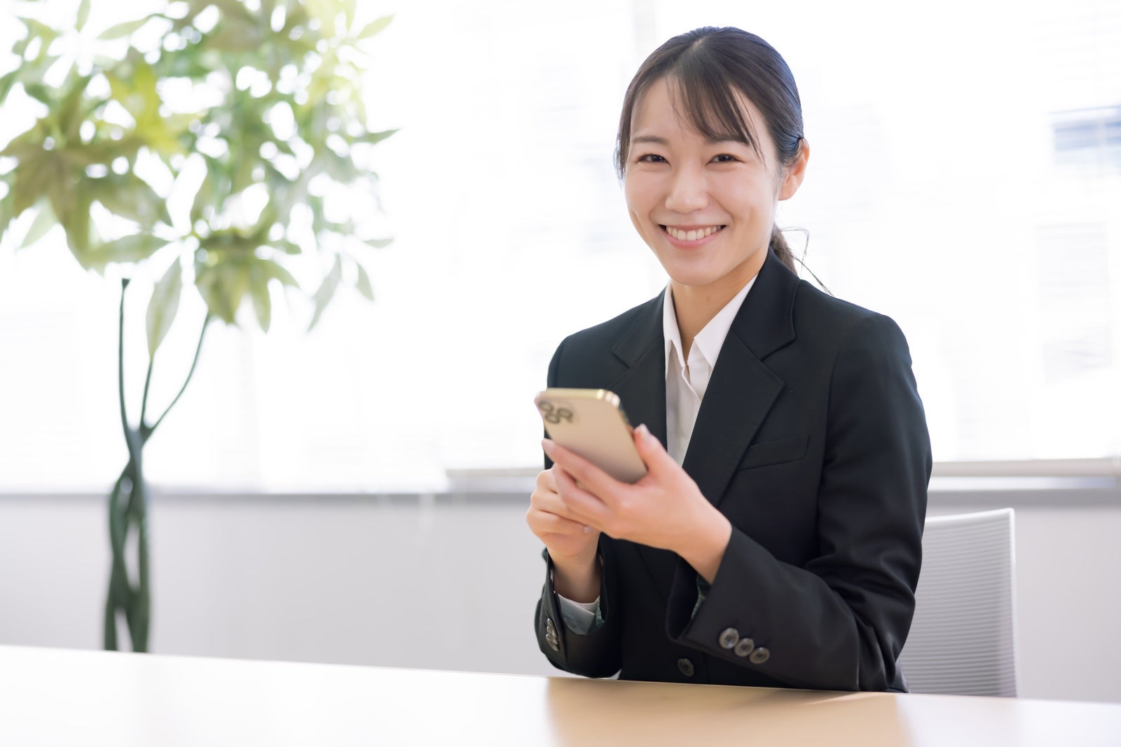黒いスーツの女性がオフィスでスマートフォンを持ち笑顔を見せている［モデル：SAKI］