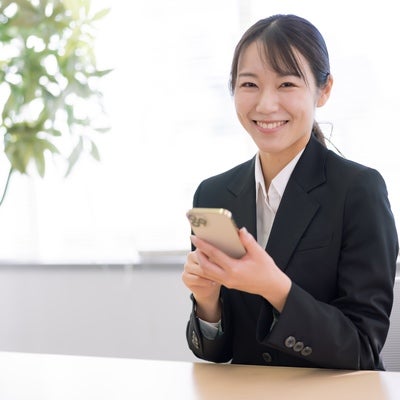 スマホを持ってにっこり笑顔の会社員女性がオフィスの机で仕事中の写真