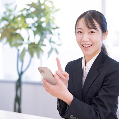 明るいオフィス空間でスマホを操作する笑顔のビジネスウーマンの写真