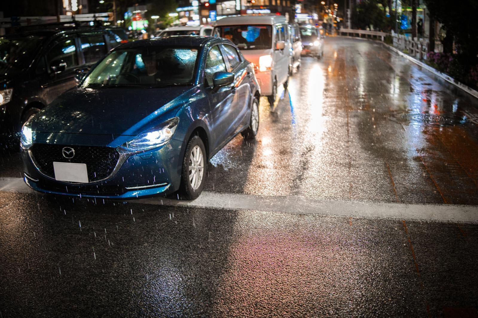 雨が降る夜の道路でヘッドライトを点けて停車する青いマツダ車と濡れた路面
