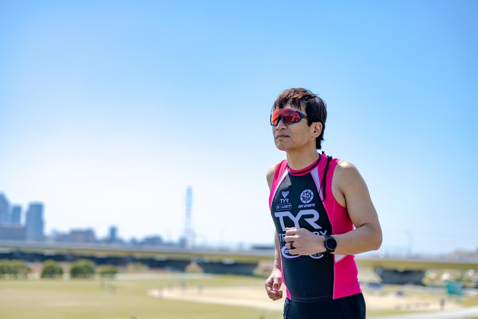 サングラスをかけピンクのウェアで河川敷を走るトライアスロン選手の男性［モデル：うえせい］