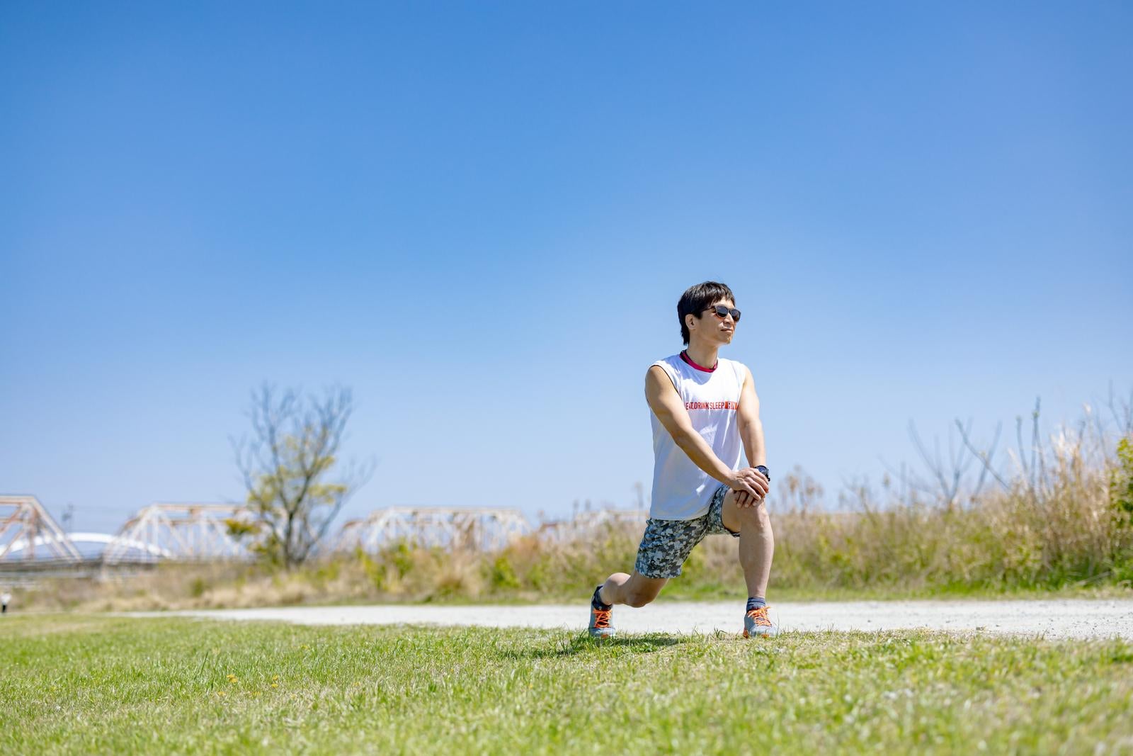 青空の下、草地で脚を伸ばしてストレッチをするタンクトップ姿の男性［モデル：うえせい］