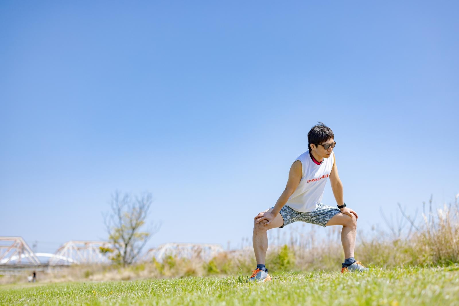 青空の下、河川敷の芝生で足を広げてストレッチをするサングラス姿の男性［モデル：うえせい］