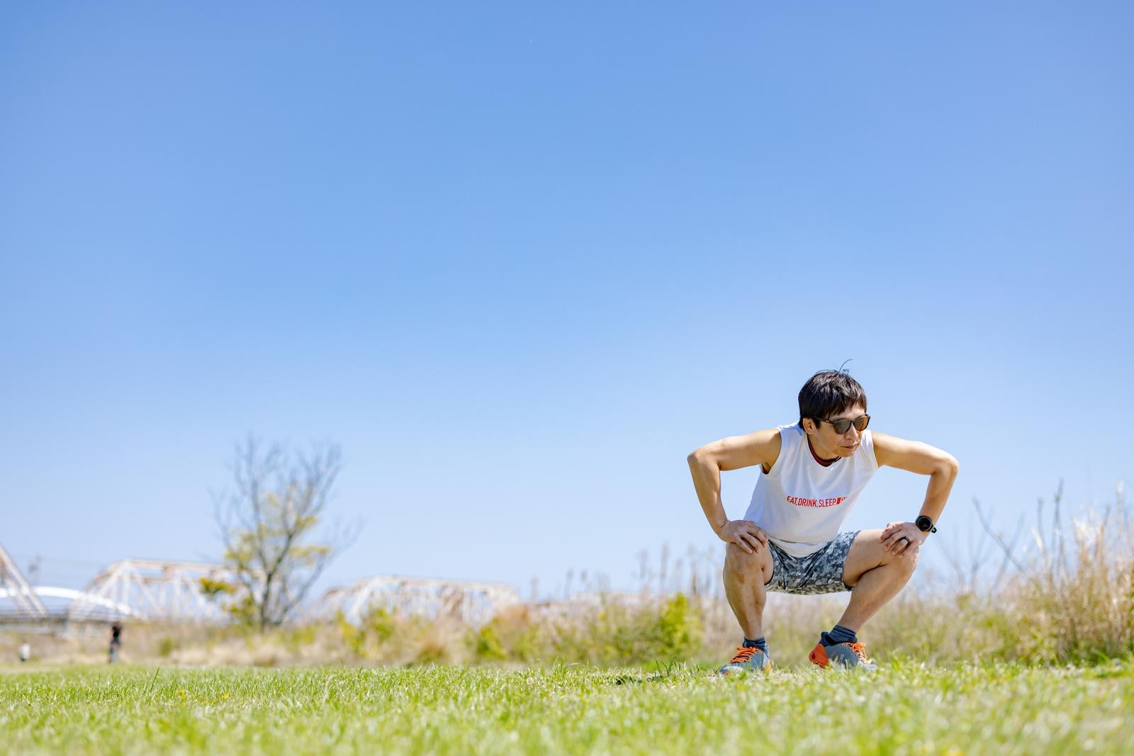 青空の下、芝生の上で屈伸ストレッチをするサングラス姿の男性ランナー［モデル：うえせい］