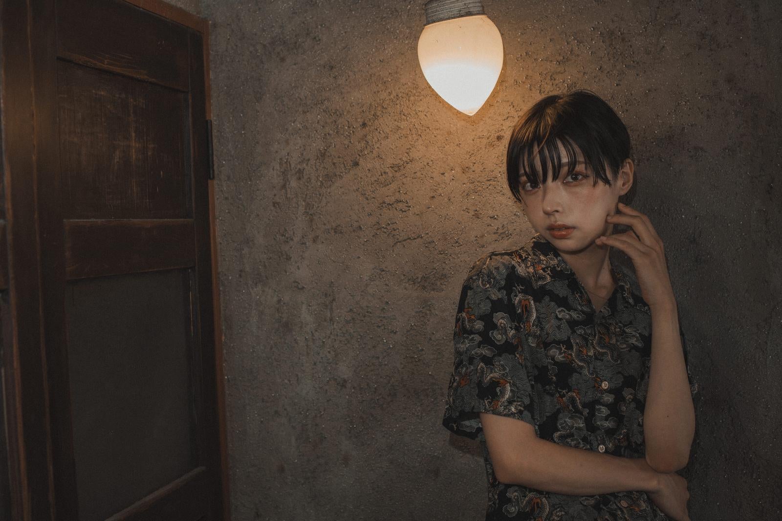 花柄シャツを着た女性が古びた壁の前で首に手を当てるポートレート写真［モデル：高尾実生］