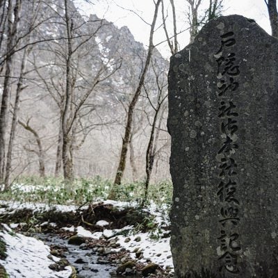 冬枯れた木々の向こうに見える戸隠山や雪の残る逆さ川を背に建つ神社御本社復興記念碑の写真