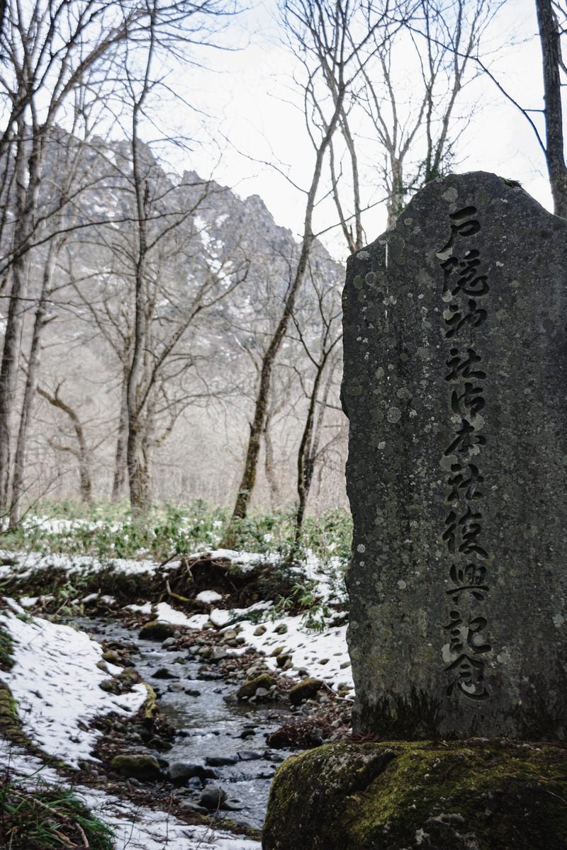 「冬枯れた木々の向こうに見える戸隠山や雪の残る逆さ川を背に建つ神社御本社復興記念碑」の写真