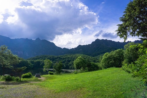 鏡池横に広がる草地と妙高戸隠連山国立公園の碑と奥にそびえる急峻な山の姿の写真