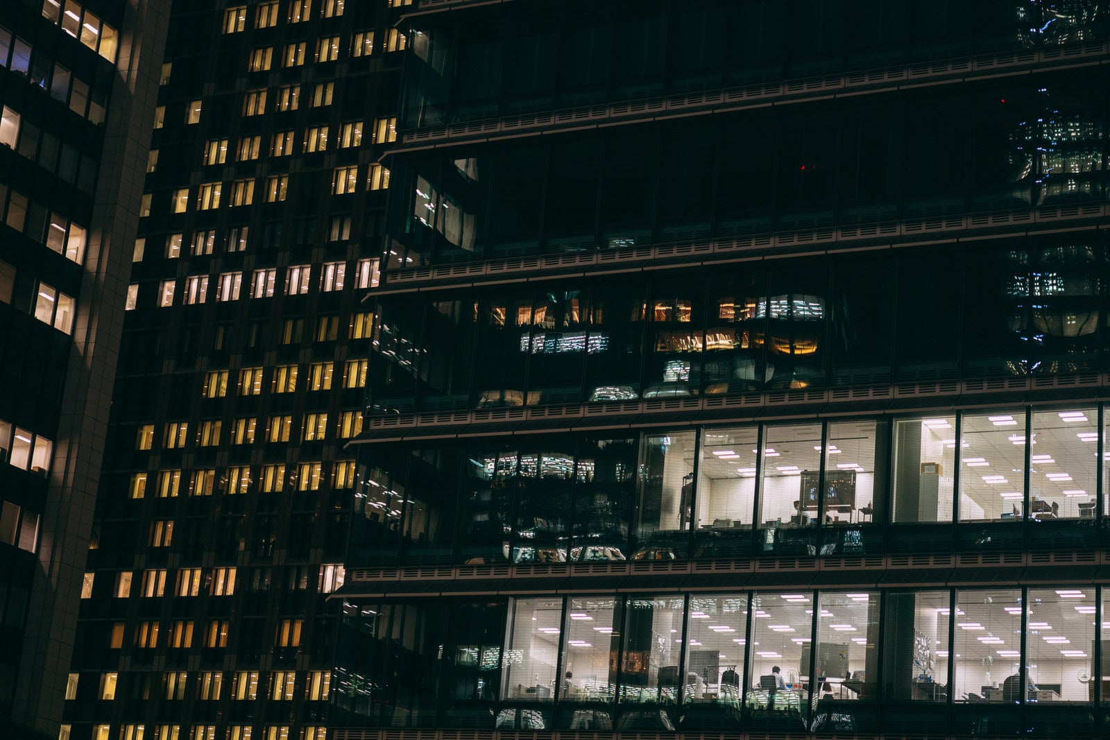 「高層ビル群の窓ガラスから見える残業中のサラリーマン」の写真