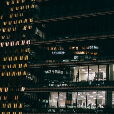 高層ビル群の窓ガラスから見える残業中のサラリーマンの写真