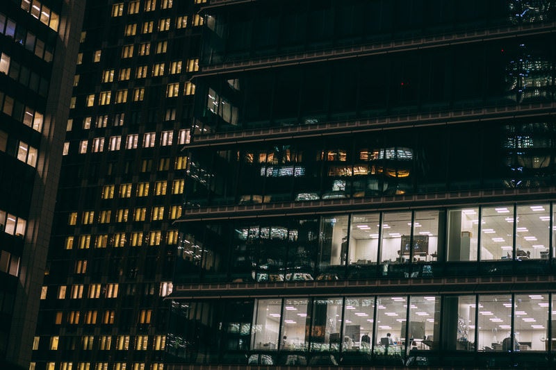 高層ビル群の窓ガラスから見える残業中のサラリーマンの写真