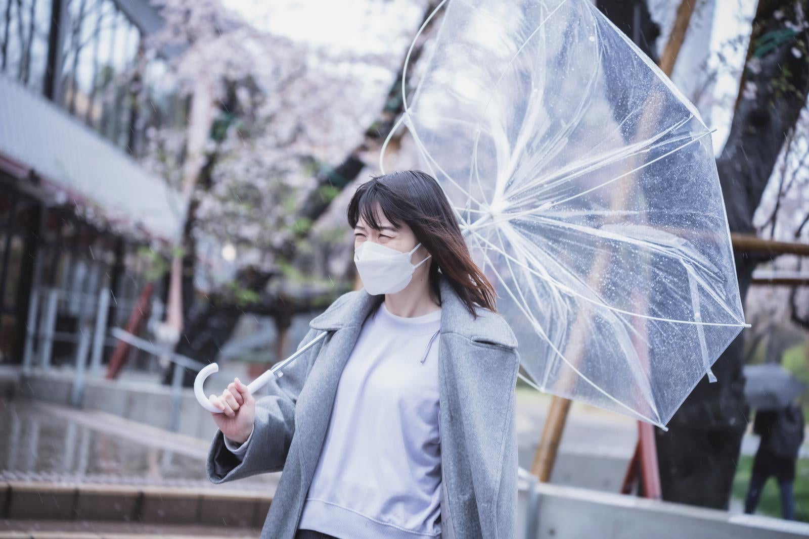台風の強風であおられた傘を持つマスク姿の女性が古い寺院前に立っている［モデル：たけべともこ］