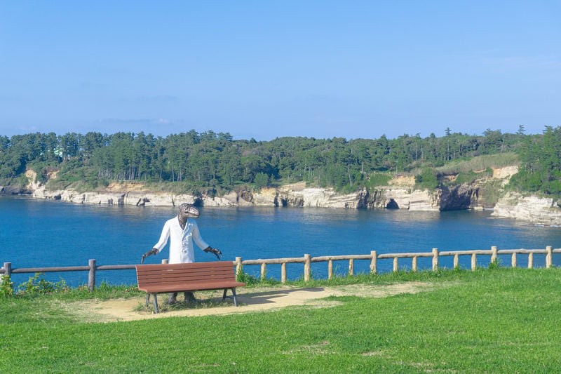 東尋坊北の芝生広場に配置された恐竜博士ベンチと福良の浜の海食崖と海食洞の写真