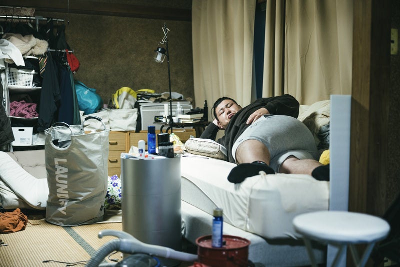 散らかった部屋で肘枕をして寝転がるデブ男性の写真
