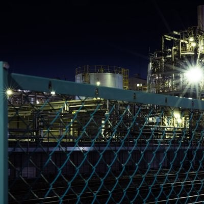 フェンス越しの工場夜景と光芒の写真