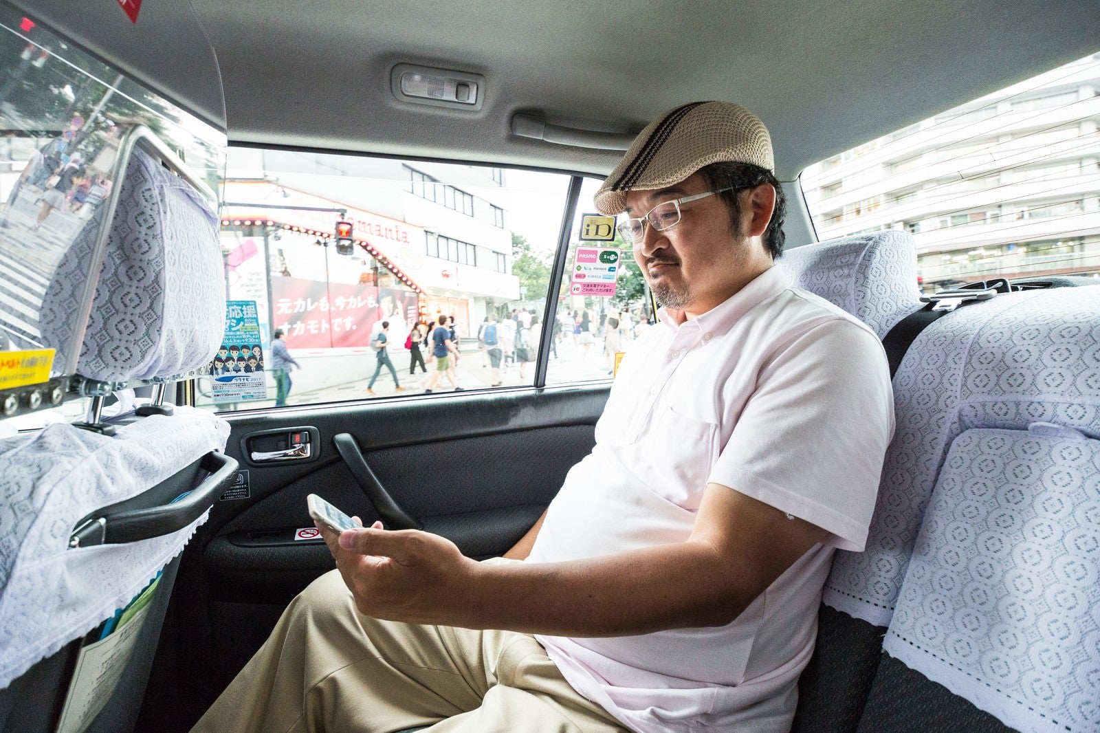 「タクシーに乗車してスマホを操作するハンチング帽のコメンテーター」の写真［モデル：ナリシゲ］