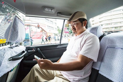 タクシーに乗車してスマホを操作するハンチング帽のコメンテーターの写真