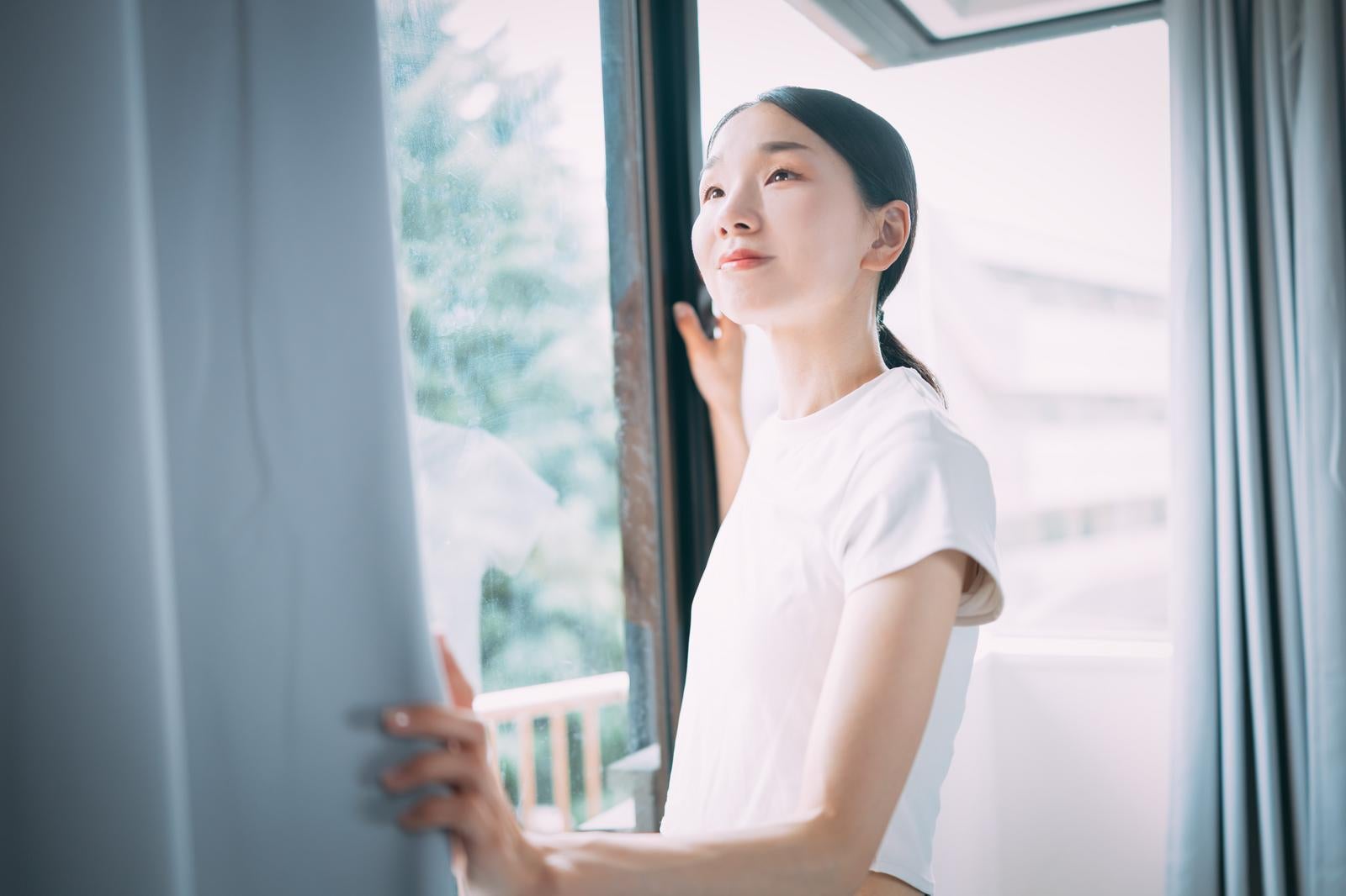 「カーテンを開けて爽やかな朝日と自然光を取り入れる女性」の写真［モデル：ひるかわえみこ］
