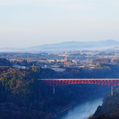 渓谷にかかる赤い大橋と秋冬の樹木、遠景の町並みの写真