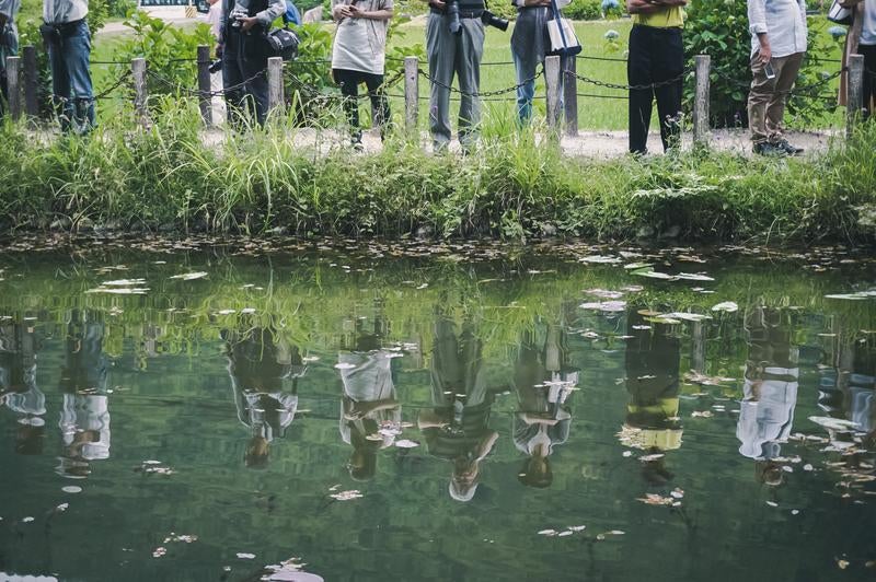 水面に反射する池の前に立つカメコを持つ観光客の姿の写真