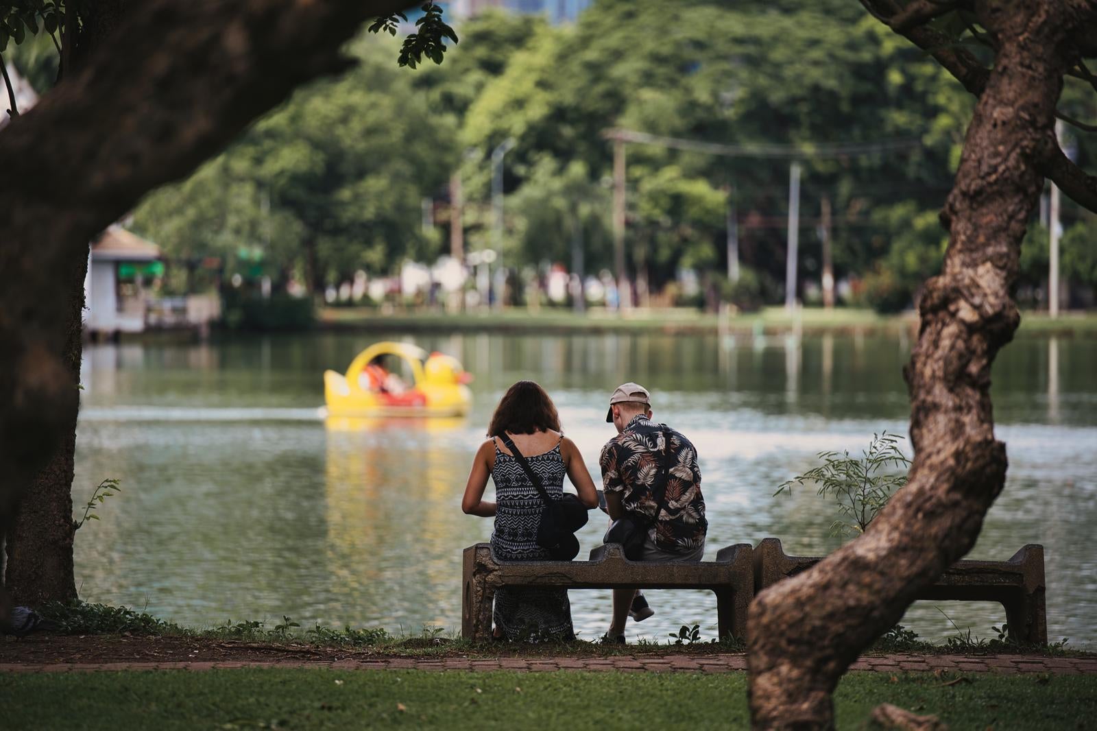 A couple sitting on a bench by the lake, with a yellow duck boat floating on the water