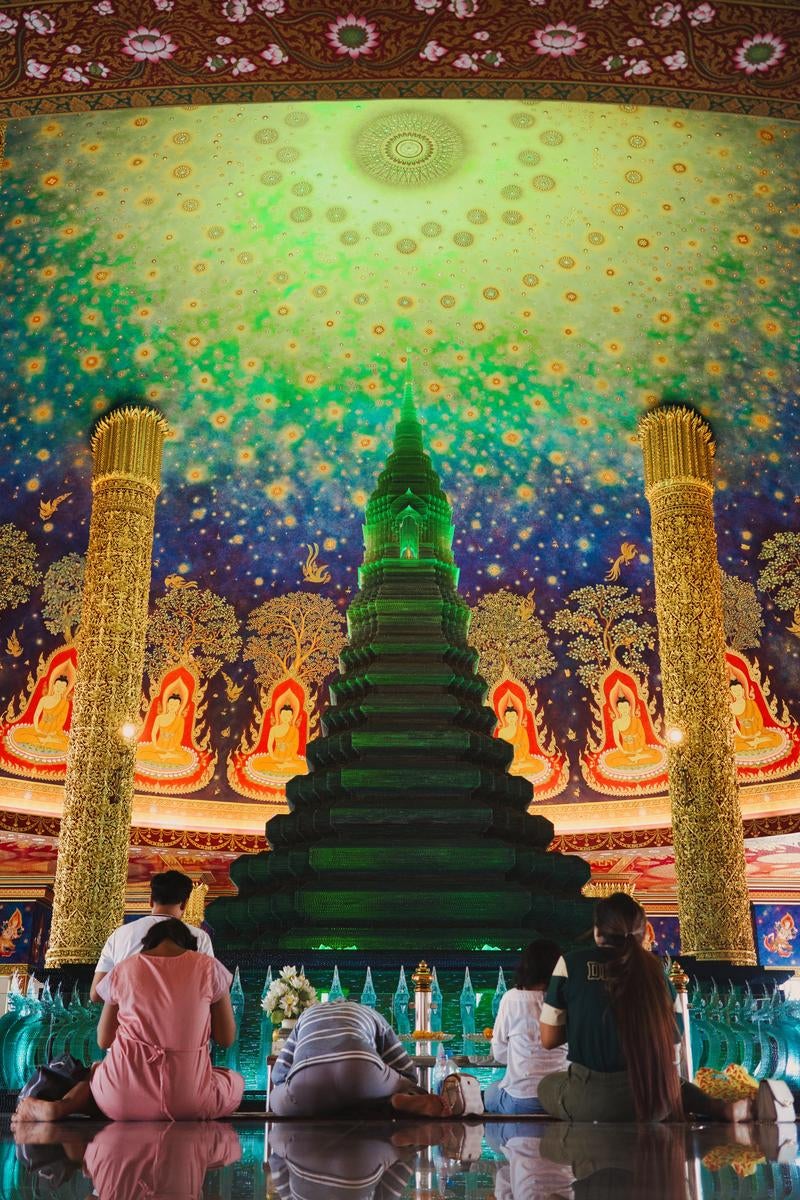 ワットパークナム寺院の装飾された天井下でエメラルド仏塔前に座り祈る参拝者たち