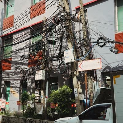 都市部の路地に立つ木製電柱に絡み合う複数の通信線と電力線の写真