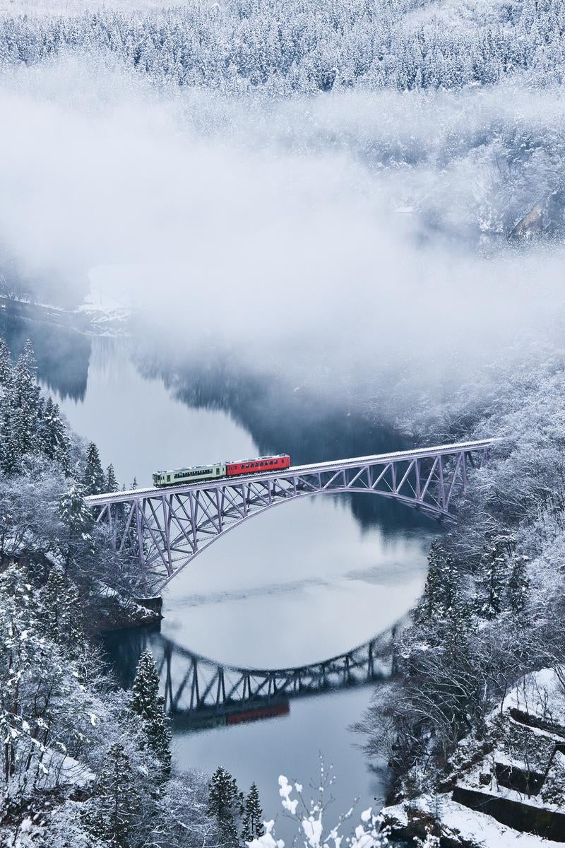 雪の積もった山々の渓谷に架かる金属トラス橋を、赤と白の列車が走行している冬景色