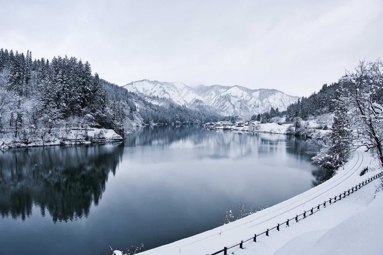 雪をかぶった針葉樹林に囲まれた川が鏡のように映り込み、遠く雪山が見える。手前には白と黒の柵が雪の上を走る冬景色