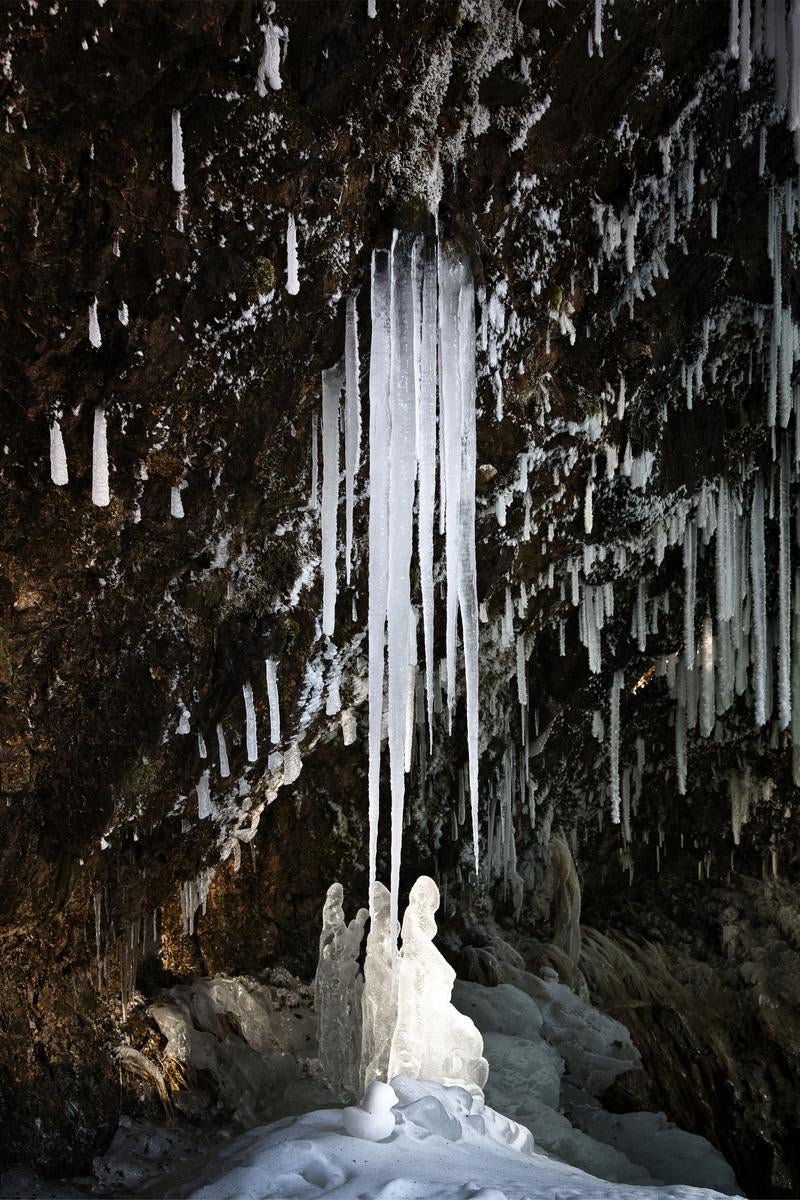 暗い岩壁の上から白い氷柱が垂直に垂れ下がり、下部に氷が盛り上がって積もっている冬の風景