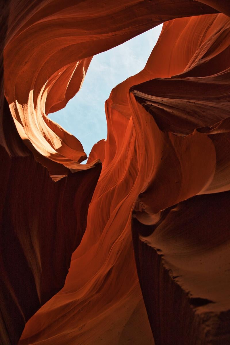 赤茶色の砂岩が波状に浸食されたアンテロープキャニオンの渓谷内部。上部から光が差し込み、空が見えている