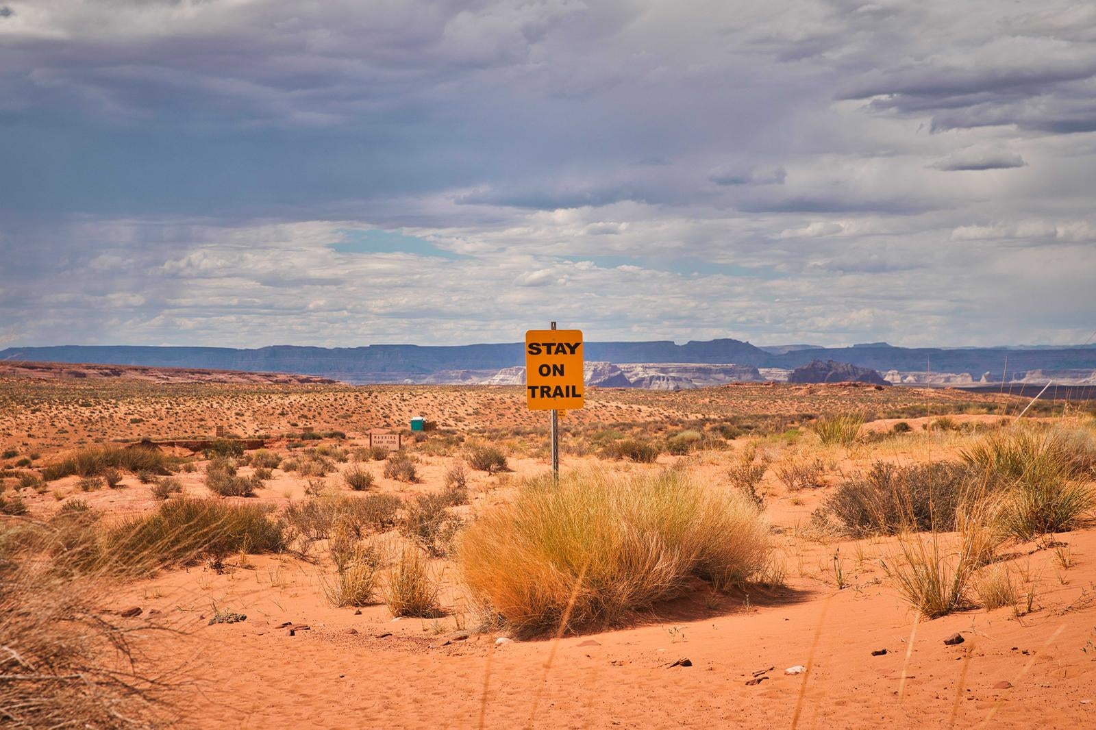 砂漠の荒野に立つオレンジ色の「STAY ON TRAIL」標識と遠くの山々