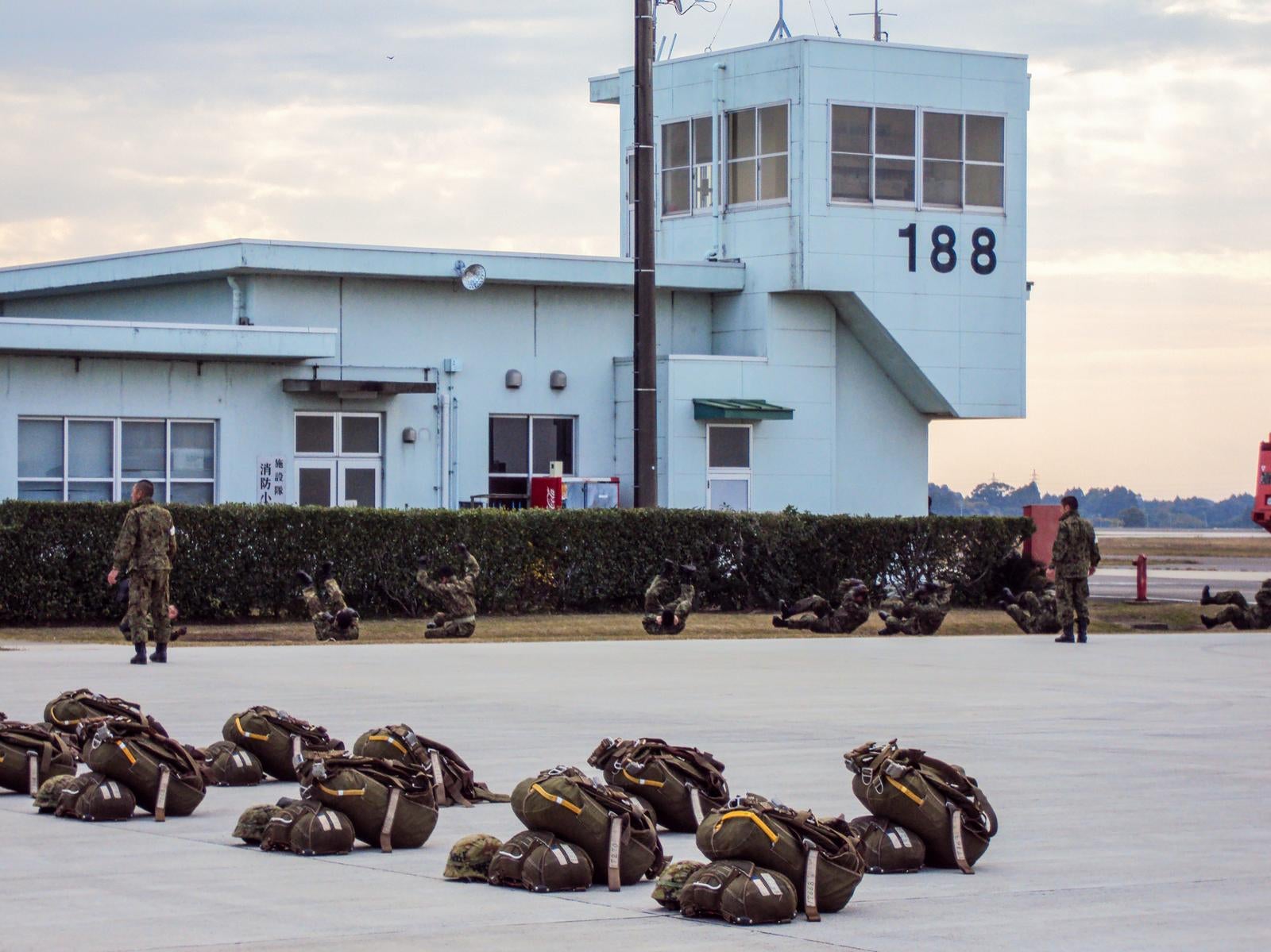 駐機場に整列して置かれた落下傘バッグと、奥で訓練する迷彩服の隊員たち