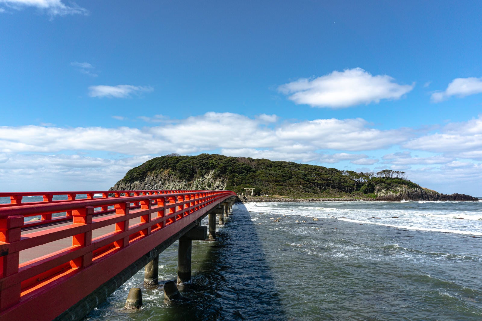 「朱色の橋の先に見える海の神様の島として崇められる雄島と寄せる波」の写真