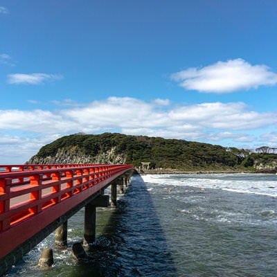 朱色の橋の先に見える海の神様の島として崇められる雄島と寄せる波の写真