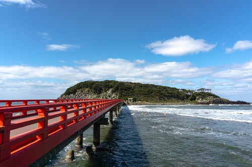 朱色の橋の先に見える海の神様の島として崇められる雄島と寄せる波の写真