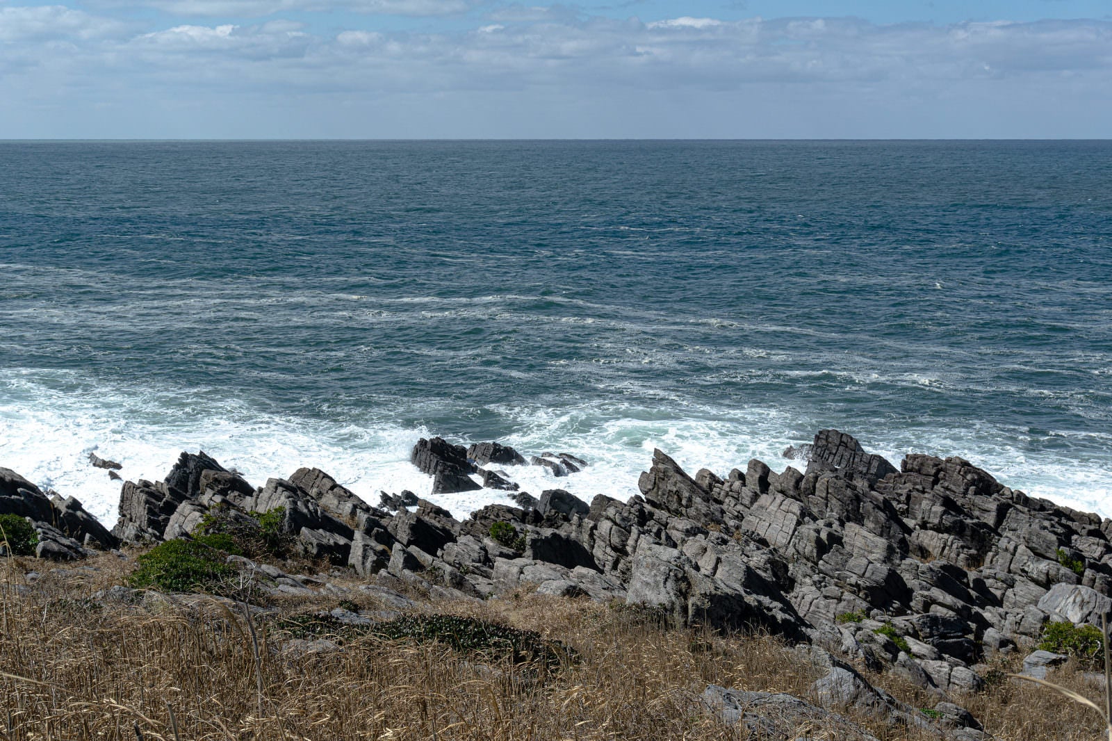 「打ち付けた波が白く泡立つ島西側岩場の波打ち際」の写真
