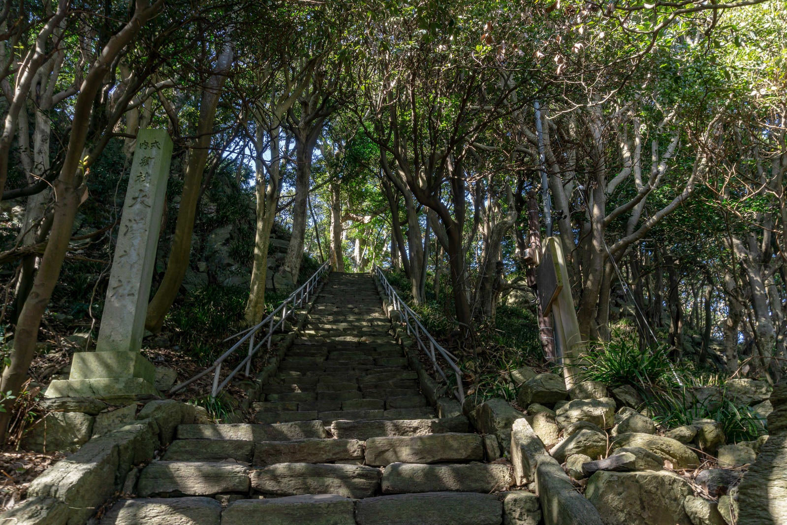 「木々に囲まれた大湊神社へと続く石段と脇に建つ社号標」の写真