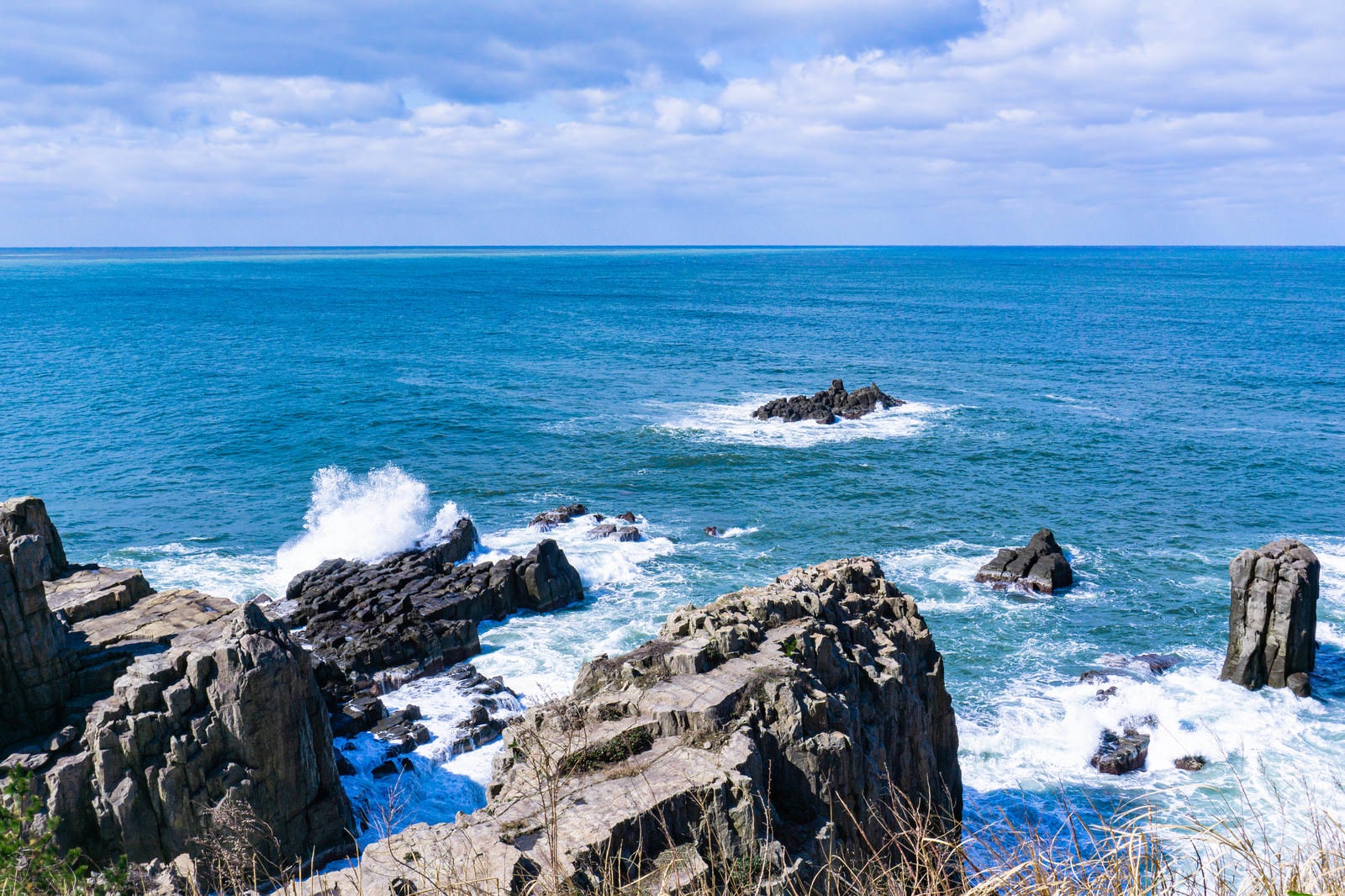 「海に浮かぶ軍艦岩と三段岩の先端で大きくあがる波しぶき」の写真