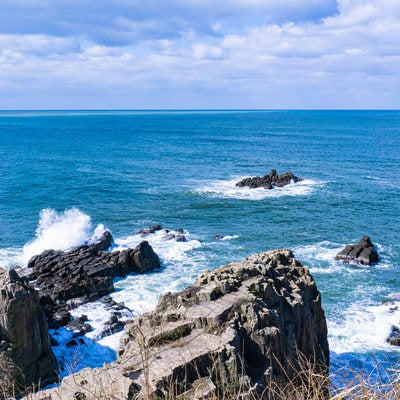 海に浮かぶ軍艦岩と三段岩の先端で大きくあがる波しぶきの写真