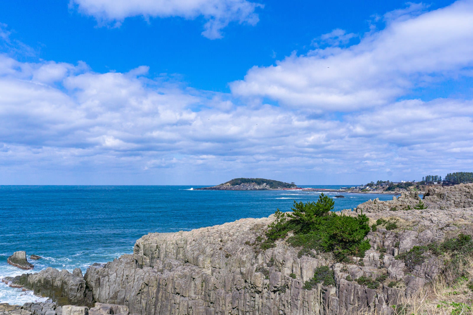 「東尋坊の切り立った崖の向うに打ち寄せる波が白く見える雄島」の写真
