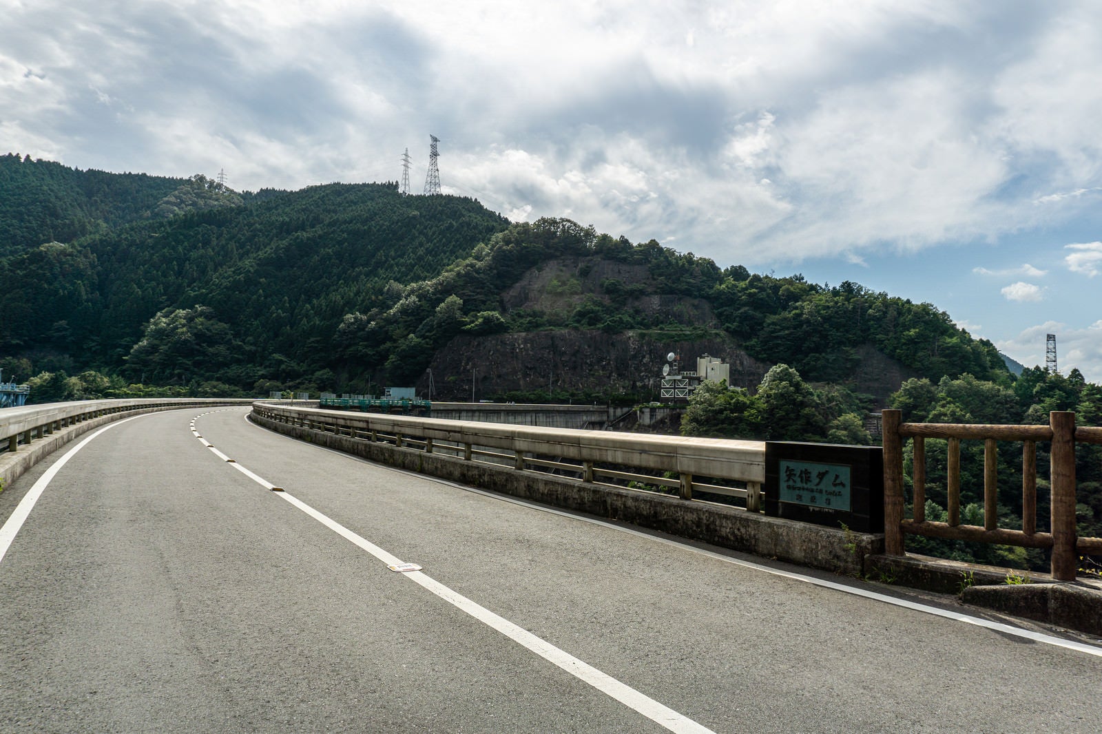 「アーチ式ダムの緩く弧を描く天端を走る県道とダム名が刻まれた親柱」の写真