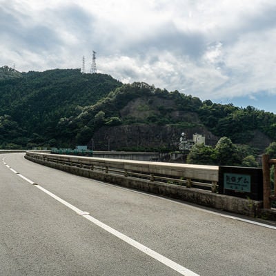 アーチ式ダムの緩く弧を描く天端を走る県道とダム名が刻まれた親柱の写真