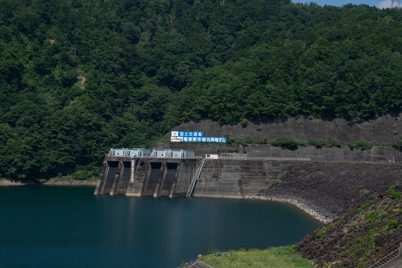 「山肌に掲げられた国土交通省とダム名の看板とその下に見える発電所取水口（福井県大野市）」の写真