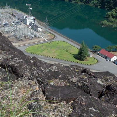 ダム天端からゴツゴツした黒い岩越しに見える長野発電所屋外送変電設備と芝生に描かれた「ONEふくい」の文字の写真
