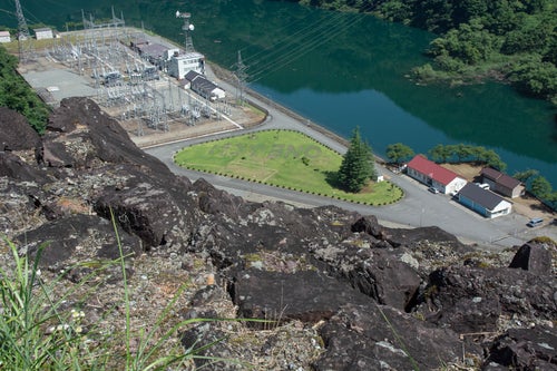 ダム天端からゴツゴツした黒い岩越しに見える長野発電所屋外送変電設備と芝生に描かれた「ONEふくい」の文字の写真