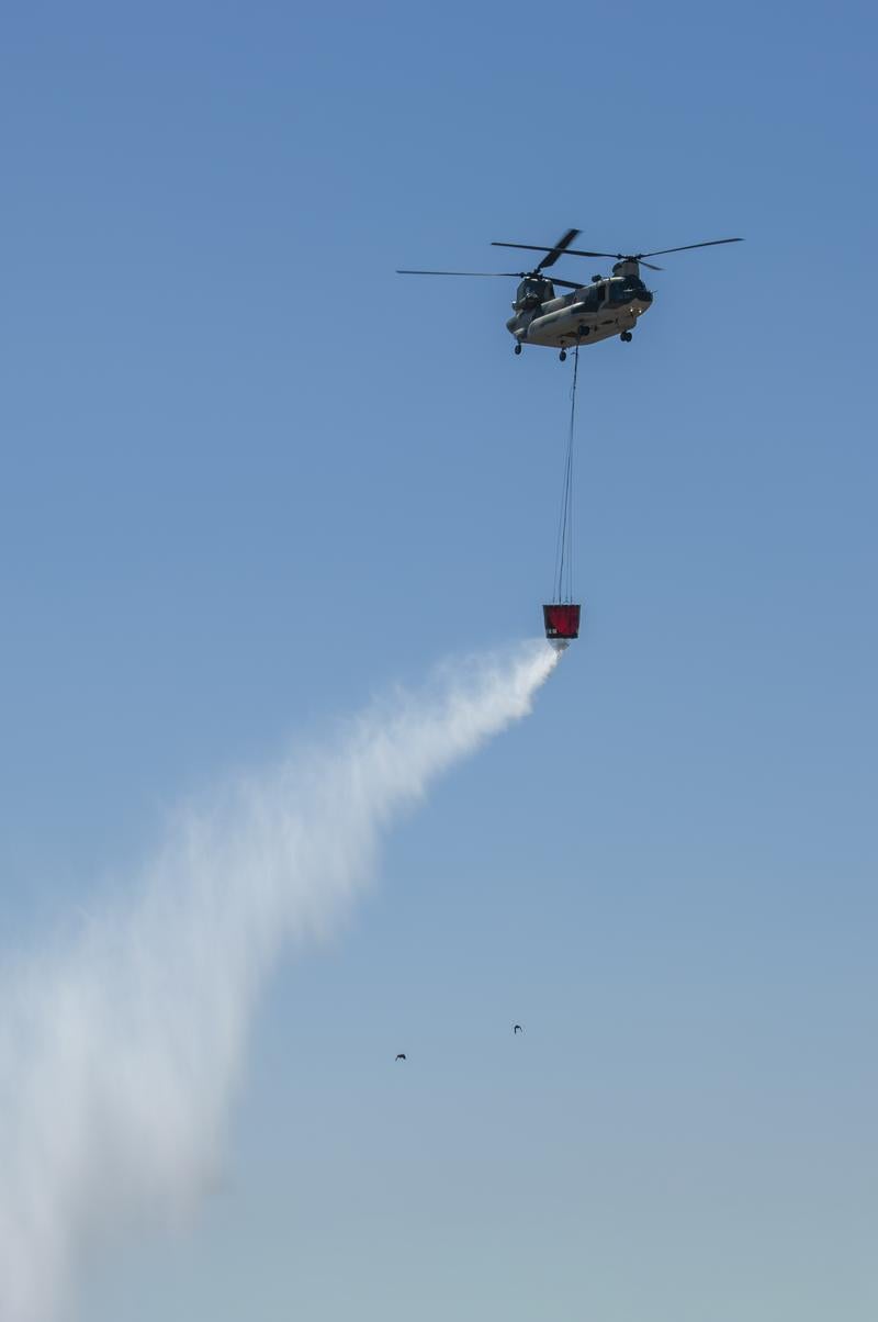青空の下で赤いバケツから放水しながら消火活動を行う自衛隊ヘリコプター